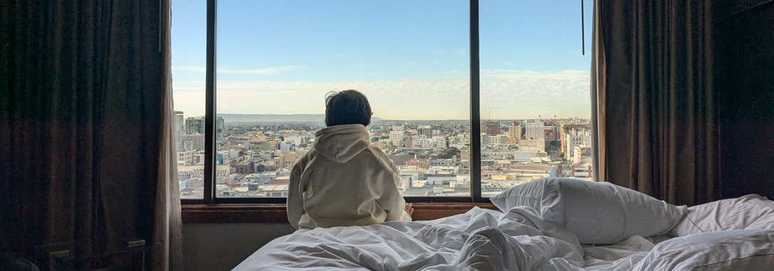A view of someone sitting on an unmade bed, looking out the window, back to the camera, with a view of buildings outside the window